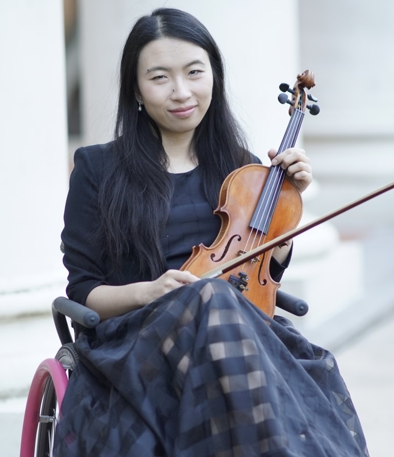 Amy Wang-Hiller seated in a wheelchair, holding a violin and bow, wearing a black dress and smiling at the camera.