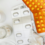 Overhead view of multiple blister packs containing different prescription medications and tablets beside medication bottles and a tissue on a white surface, representing the complexity and emotional burden of starting and managing medications with chronic illness.