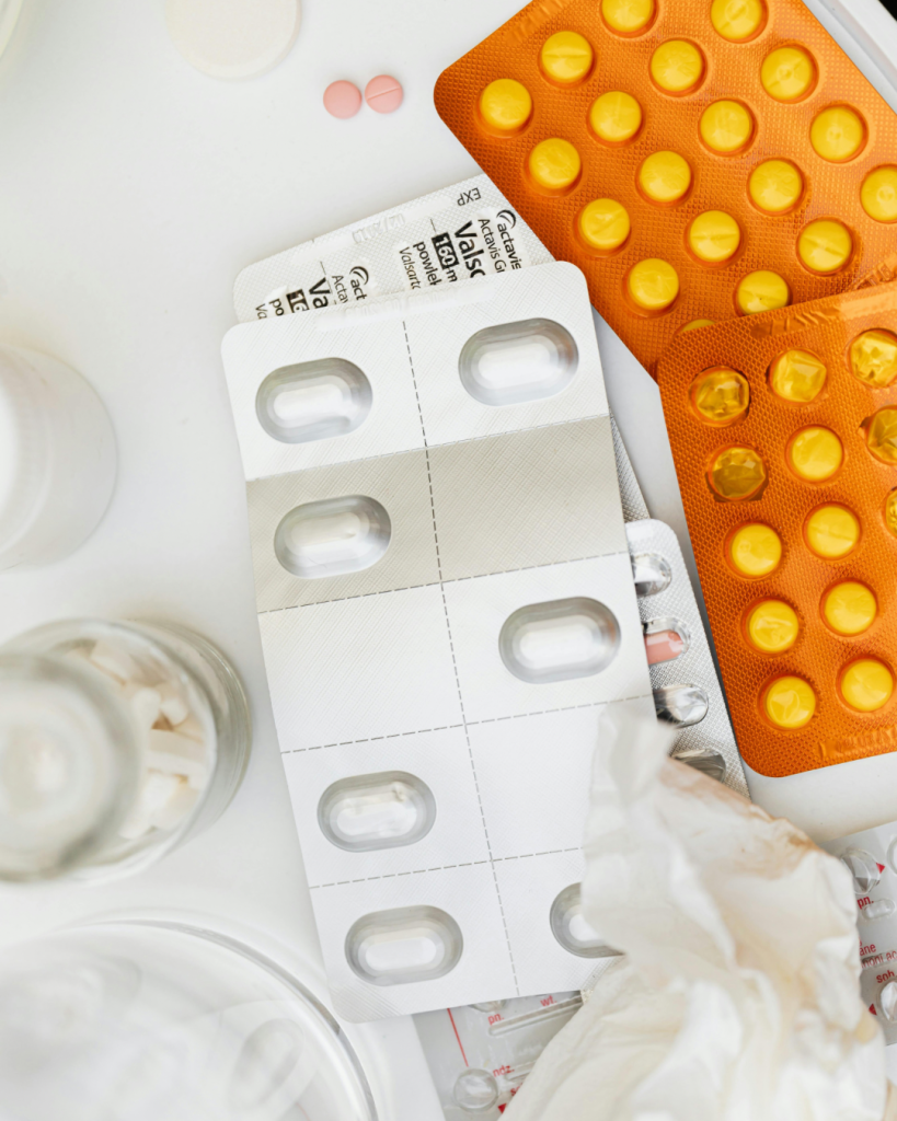 Overhead view of multiple blister packs containing different prescription medications and tablets beside medication bottles and a tissue on a white surface, representing the complexity and emotional burden of starting and managing medications with chronic illness.