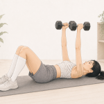 A minimalist watercolor-style illustration of a young Asian woman performing a dumbbell chest press while lying on an exercise mat. She wears a light zebra-print athletic top, gray shorts, white compression socks, and white trainers. Beige kinesiology tape is applied to her upper arm. The bright, neutral-toned home workout space includes a potted plant and simple shelving, conveying a calm, low-impact strength training environment.