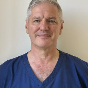 Head-and-shoulders portrait of Professor Mark S. Whiteley wearing navy blue medical scrubs embroidered with "The Whiteley Clinic – Pioneering Vein Treatments." He has short gray hair and a slight smile and is photographed against a plain light-colored background.