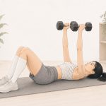 Illustration of a woman with EDS lying on a gray exercise mat in a bright, neutral-toned home workout space. Wearing a striped crop top, gray shorts, knee-high white socks, and sneakers, she holds two dumbbells above her chest. Kinesiology tape is wrapped around one elbow, with potted plants, folded towels, and storage baskets in the background.