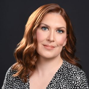 Headshot of Allison Wallis, smiling, with shoulder-length red hair against a dark background.