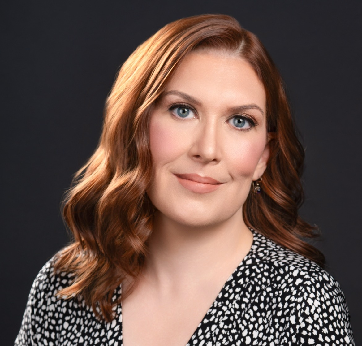 Headshot of Allison Wallis, smiling, with shoulder-length red hair against a dark background.